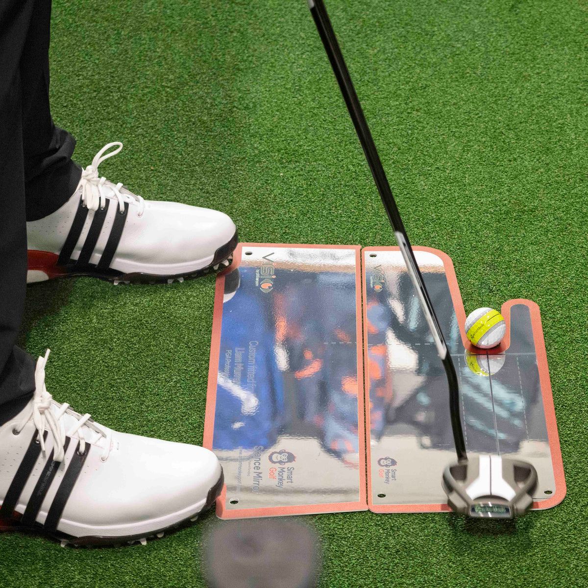 Golfer at address over a Club Stance Mirror on a practice green, putter behind the ball, showing the mirror's size relative to a golfer's stance